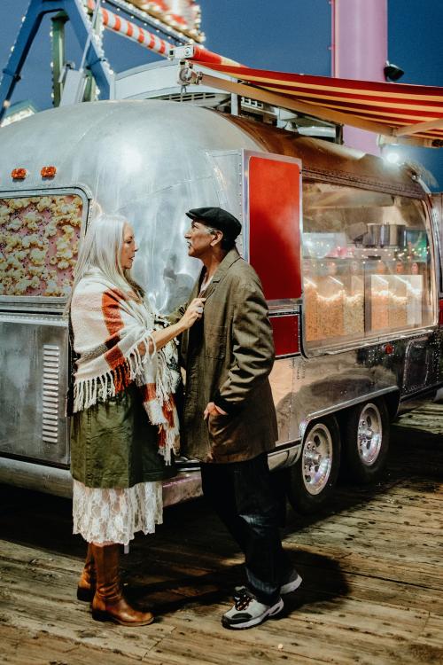 Cool senior couple standing by the food truck inside of an amusement park - 1202584